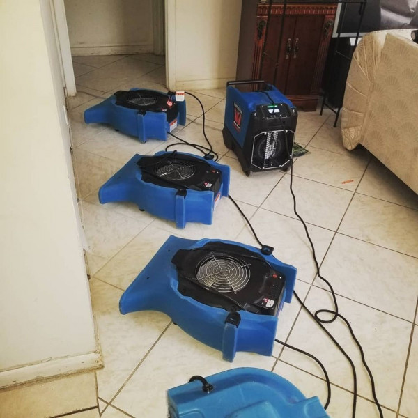 Three blue air movers and a dehumidifier running on a tile floor in a hallway after a leak