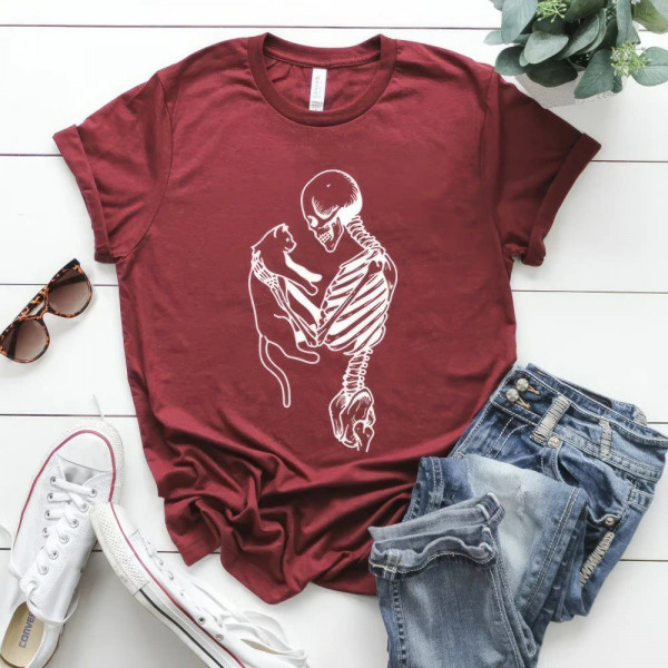 Maroon T-shirt with a white print of a skeleton holding a cat, laid out with jeans and sneakers