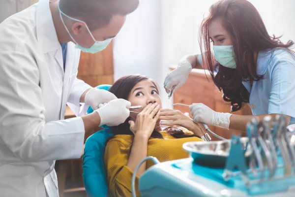 Rädd kvinna i tandläkarstol omgiven av tandläkare och tandsköterska